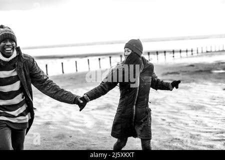 Playful couple in warm clothing holding hands on winter beach Stock Photo