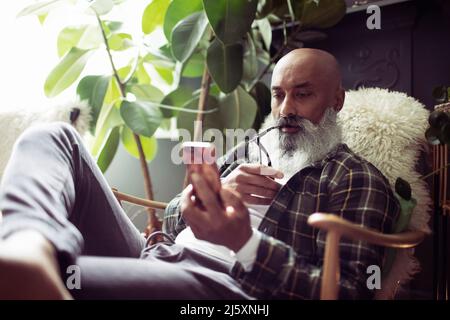 Mature man with beard using smart phone in armchair Stock Photo