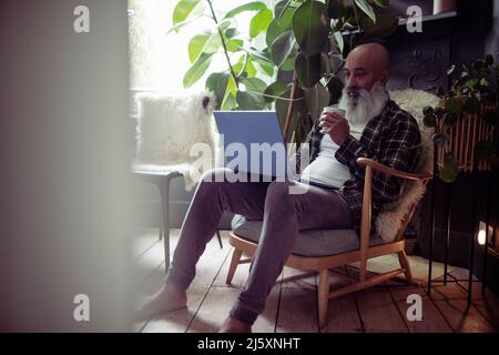 Mature man with tea using laptop in armchair at home Stock Photo