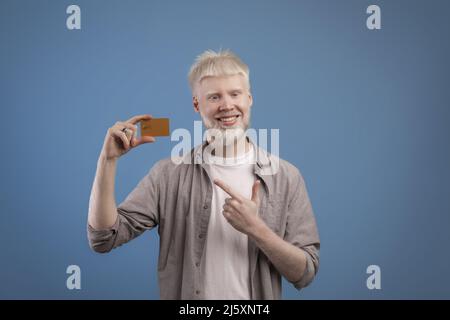 Excited albino guy pointing at bank card in his hand and smiling at camera, recommending bank over blue background Stock Photo