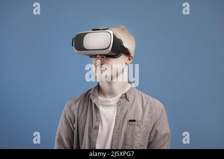 Virtual gadgets for entertainment. Young male student using vr glasses and looking at empty space, blue background Stock Photo
