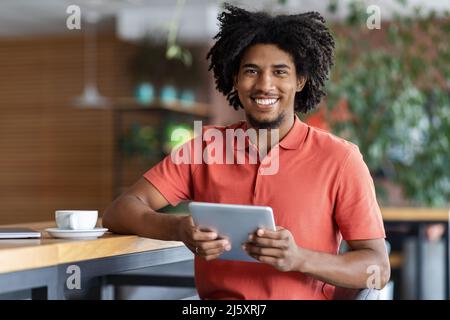Cheerful millennial black curly man blogger typing on tablet at table with cup of coffee in modern cafe interior Stock Photo