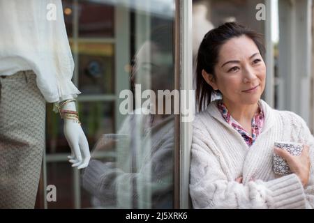 Smiling thoughtful female shop owner with coffee outside shop Stock Photo