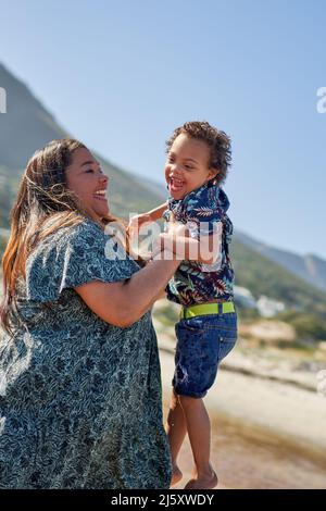 Happy mother lifting cute son with Down Syndrome on beach Stock Photo