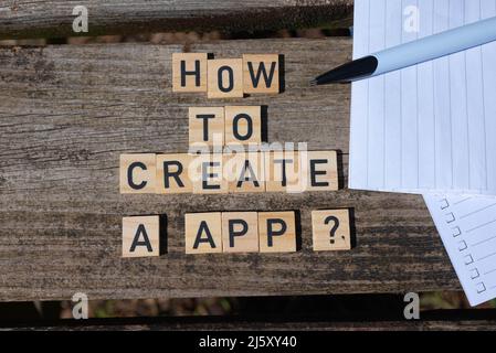 How to create a app? Black capital letter words on wooden toy blocks on a natural garden table background with paper and pencil. Perfect for websites. Stock Photo