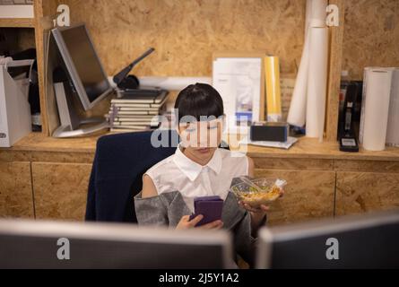 Businesswoman eating lunch and using smart phone in office Stock Photo