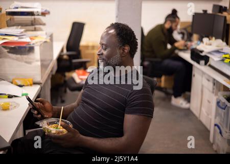 Businessman eating lunch and using smart phone at desk Stock Photo