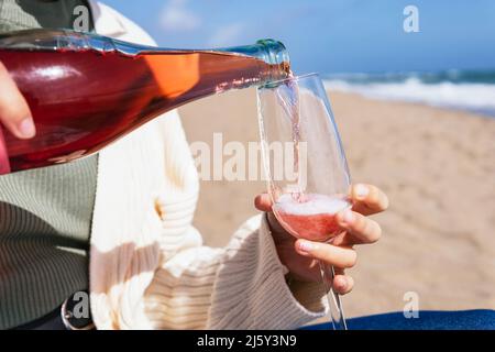 Crop anonymous female pouring rose sparkling wine from bottle into glass while sitting on sandy shore near waving sea in tropical resort Stock Photo