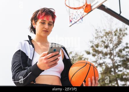 Low angle of concentrated lady with dyed hair holding basketball and browsing mobile phone on outdoor sports ground Stock Photo