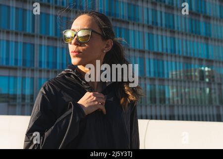Fit female athlete in black activewear and sunglasses standing looking away while having break during training on sunny street in city Stock Photo