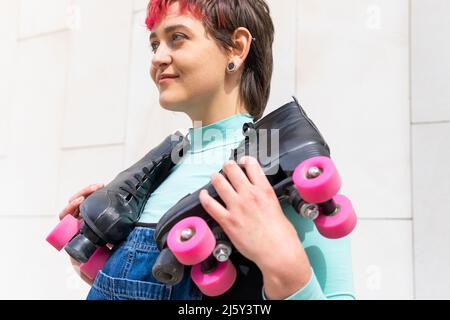 Side view of short haired Woman in denim overall holding roller skates while walking near tiled wall on street Stock Photo