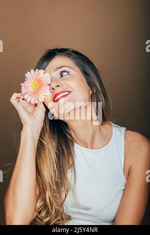 Peaceful female model with red lips covering eye with gentle pink gerbera bloom in studio against beige background Stock Photo