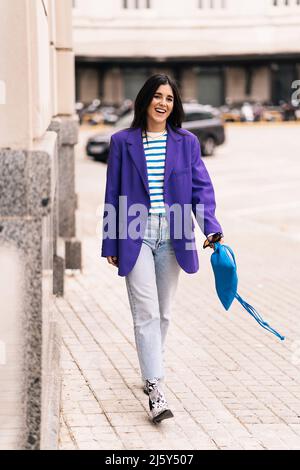 Full body of positive female with black hair in stylish outfit strolling on paved walkway along building on street of city Stock Photo