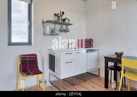 Interior of kitchen with various utensils and red microwave oven on white cabinet placed near glass door overlooking backyard with building Stock Photo