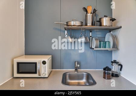 Shelves with various utensils and dishware hanging on wall above sink and white microwave oven in light kitchen of apartment Stock Photo