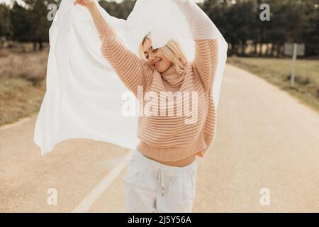 Woman in pink sweater standing with raised arm on asphalt road in countryside in daylight with white cloth covering head Stock Photo