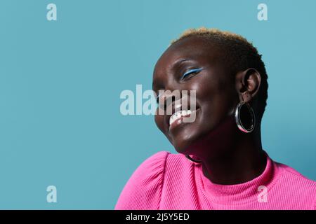 Positive African Woman with bright makeup and short dyed hair looking up with closed eyes while standing on red background in light studio Stock Photo
