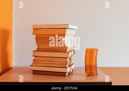 Pile of books on a piece of furniture next to a glass of transparent glass of orange color Stock Photo