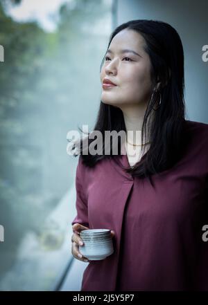 Thoughtful young woman drinking tea at window Stock Photo