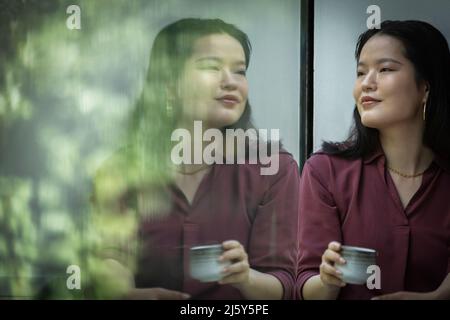 Thoughtful young woman drinking tea at window Stock Photo