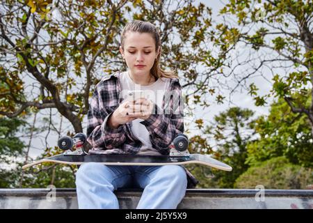 Young female with black skateboard browsing on smartphone while sitting in skate park Stock Photo