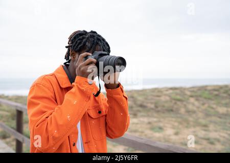 Focused African American male photographer taking picture on professional photo camera while standing on coast near sea Stock Photo