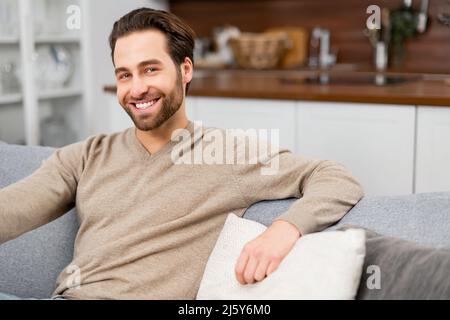 Handsome optimistic mature man staring at the camera with cheerful toothy smile. Happy guy in smart casual wear sitting on the sofa at home. Lifestyle and wellbeing concept Stock Photo