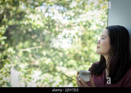 Thoughtful young woman drinking tea and looking out window Stock Photo