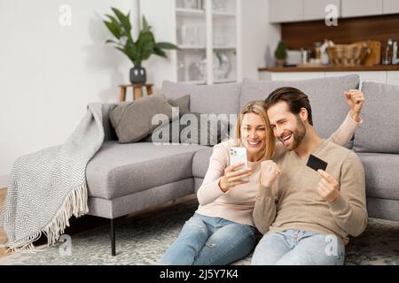 Overjoyed caucasian couple shopping together online. Excited woman and man holding smartphone and credit card, making purchases online, order, buying something in e-shop Stock Photo