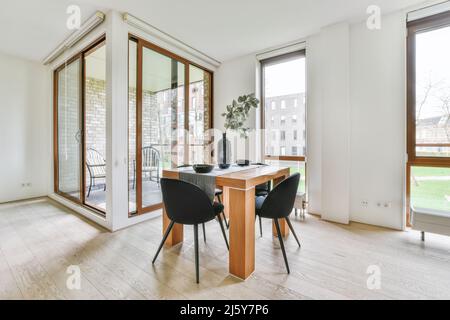 Chairs placed at square table with tableware and plant in vase in light spacious dining room with windows and glass doors in apartment Stock Photo