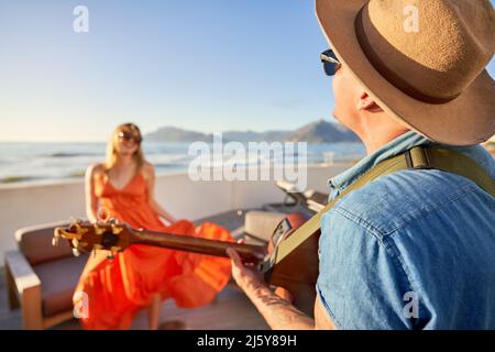 Happy couple playing guitar and dancing on sunny patio with ocean view Stock Photo