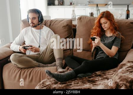 Full body of man in headphones playing video game near redheaded girlfriend browsing smartphone while sitting together on sofa in living room Stock Photo