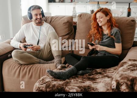 Full body of happy man in headphones playing video game near redheaded girlfriend browsing smartphone while sitting together on sofa in living room Stock Photo