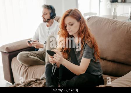 man in headphones playing video game near redheaded girlfriend browsing smartphone while sitting together on sofa in living room Stock Photo