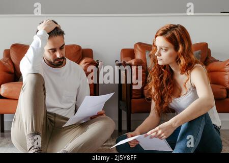 Serious couple reading documents while sitting together on floor near armchairs in light living room at home Stock Photo