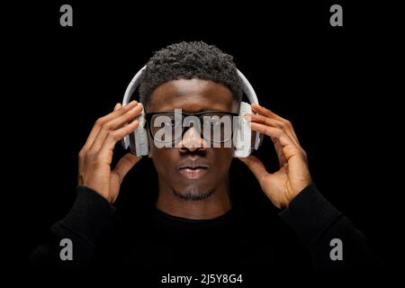 Young African American man in glasses touching in wireless headphones on head and looking at camera against black background Stock Photo
