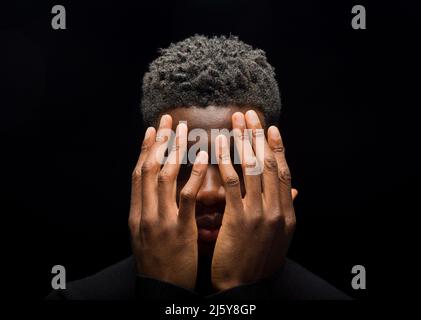 Head of upset black man with short curly hair covering face with hands on dark background in studio Stock Photo