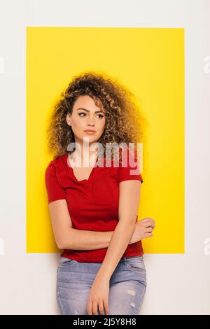 Young curly haired Woman in red t-shirt looking away on yellow and white background Stock Photo