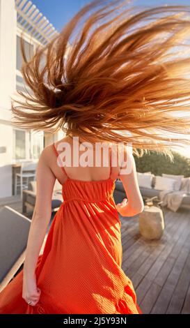 Carefree woman in orange dress flipping long red hair on sunny patio Stock Photo