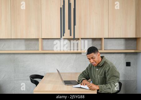 Concentrated positive hispanic man thinking, taking notes while sitting at table with laptop and smartphone at home. Author writing an idea for his book, study online. E-learning concept Stock Photo