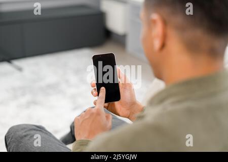 Back view of the focused man sitting on the sofa at home and holding smartphone. Hispanic guy reading news, spend leisure time in social networks, dark-haired male texting and chatting online Stock Photo