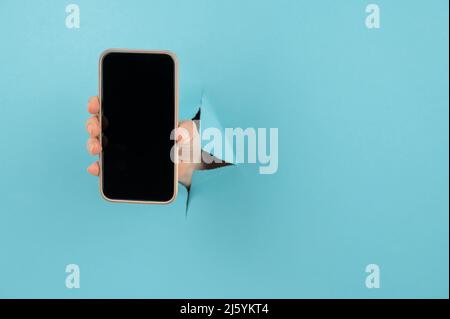 A female hand sticking out of a hole from a blue background holds a smartphone with a black screen.  Stock Photo