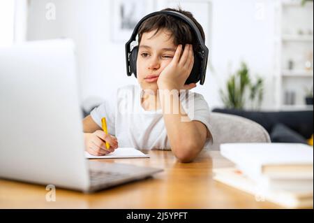 Tired schoolboy doing homework and listening music in the headphones, studying on the distance from home, using trendy laptop, preteen boy writing tasks in the notebook. Online education concept Stock Photo