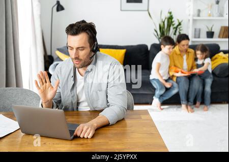 Busy parent is working on the distance. Man using laptop for freelance work sitting at the table at home while family sitting peacefully together on the sofa Stock Photo