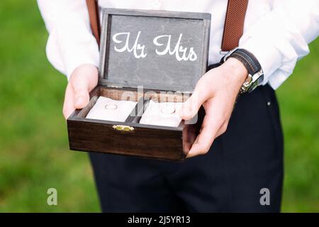 Best man holds wooden box with golden wedding rings on white pillows. Outdoor summer wedding ceremony. Rustic wedding. Mr and Mrs. Stock Photo