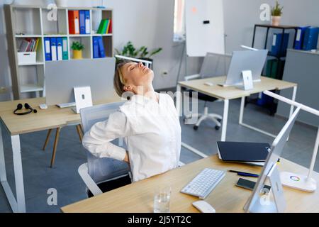 Mature business woman sits at her workplace in the office and holds on to her lower back. Health problems due to sedentary work Stock Photo