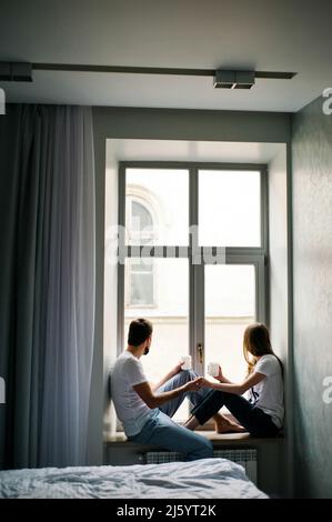 Rainy morning at home. Happy young man and woman sitting on windowsill with cups of coffee. Isolation, quarantine. Stock Photo