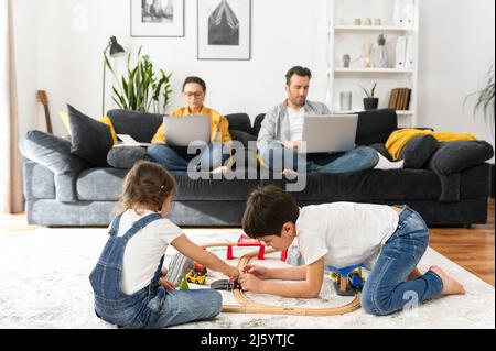 Father and mother working on the distance during quarantine. Parents work remotely with laptops sitting on the couch in living room, two sweet kids playing with toys on the foreground Stock Photo