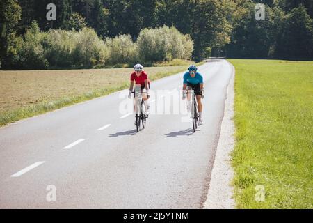 Front view of a woman and man, professional racing bikers in sportswear, riding along an asphalt road in the beautiful green nature. Stock Photo