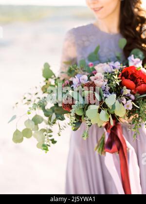 Beautiful smiling bride holds large purple wedding bouquet with red flowers and eucalyptus leaves at the sand beach. Violet wedding dress. Silk and la Stock Photo